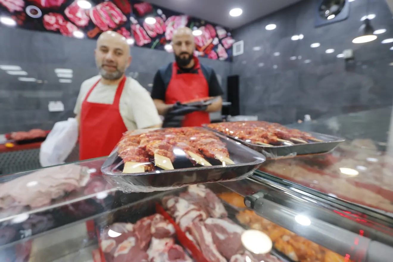 Tabletts mit frisch geformten Köfte und Spießen auf Holzstäbchen über der Fleischtheke, dahinter zwei Metzger in roten Schürzen